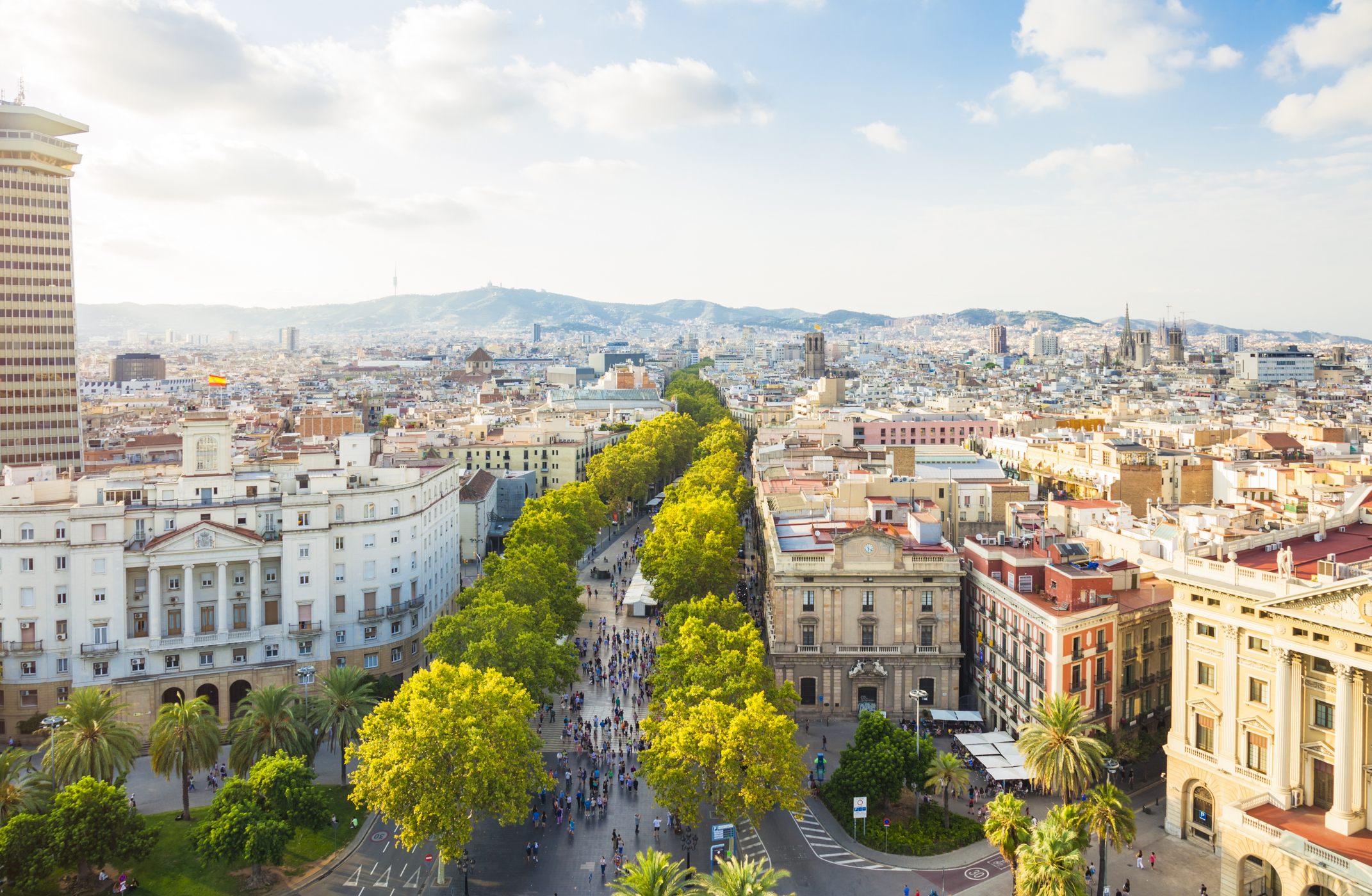 Aerial view of a tree-lined boulevard in Barcelona with pedestrians, historic buildings, and the city skyline under a bright sky.