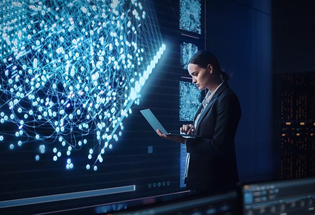 Professional reviewing data on a laptop while standing beside a large digital network visualization in a dark control room.